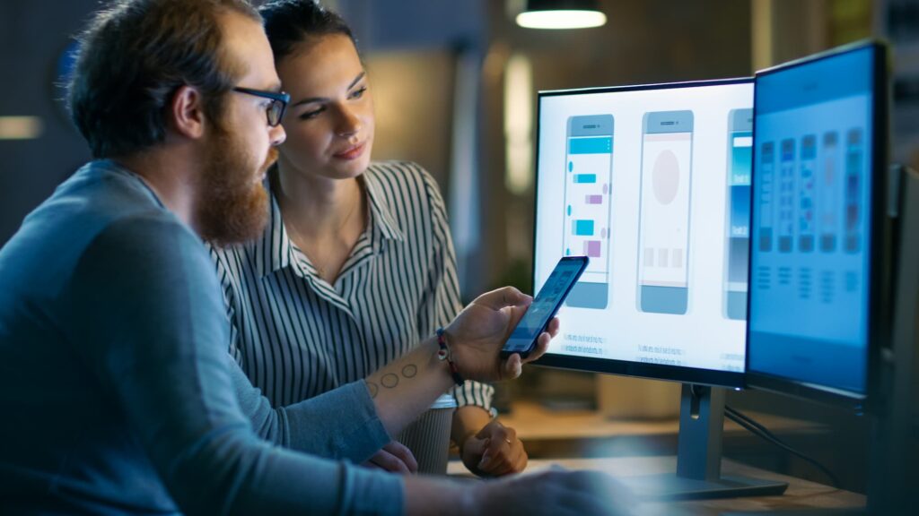 Mobile app development - A man and women review mobile app designs on a computer screen; one holds a smartphone.

