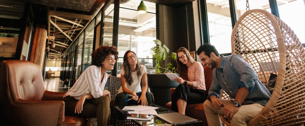 Business intelligence - Four people in an office lounge discussing around a glass table with laptops and papers.