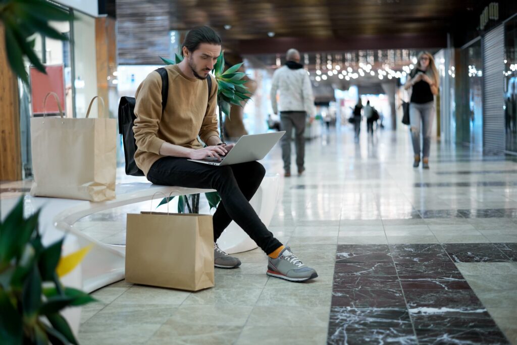 Commercial mix modelling - Young man in a shopping centre sitting on a bench with his laptop on his lap.